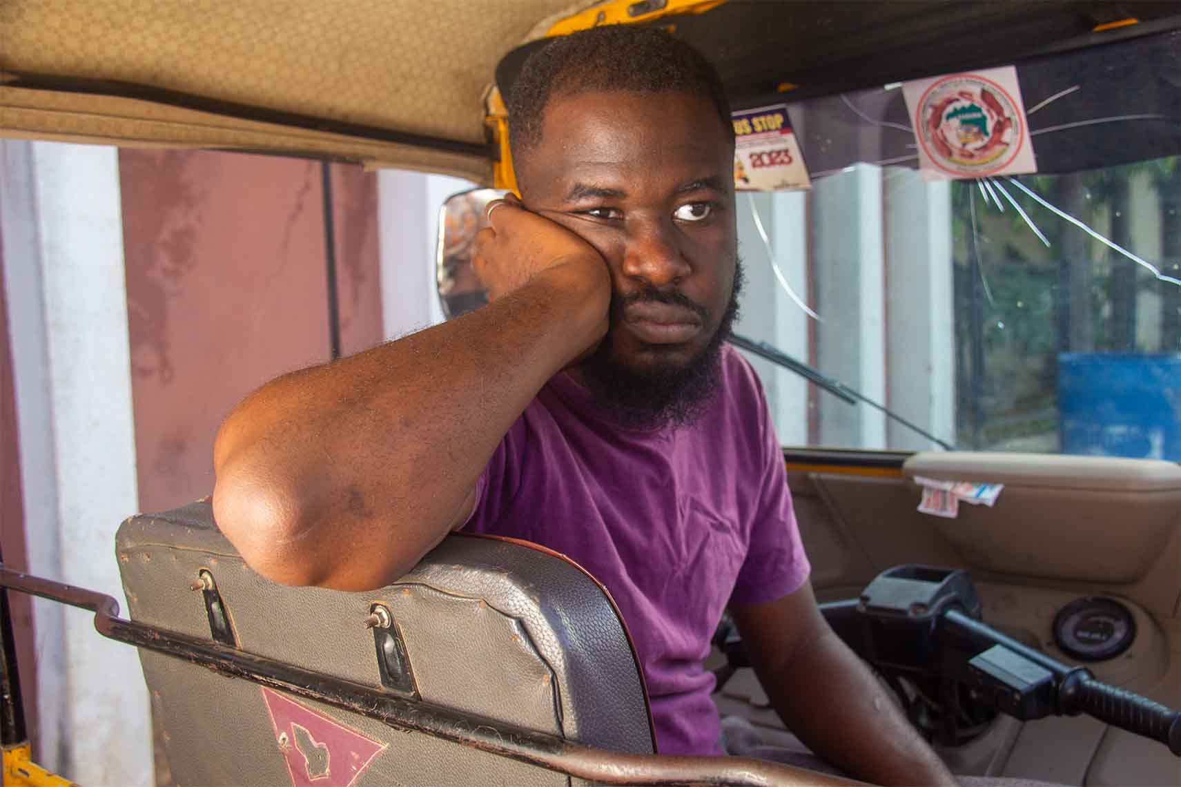 An African tricycle rider sitting inside his tricycle with his hand on his cheek, looking sad and distressed.